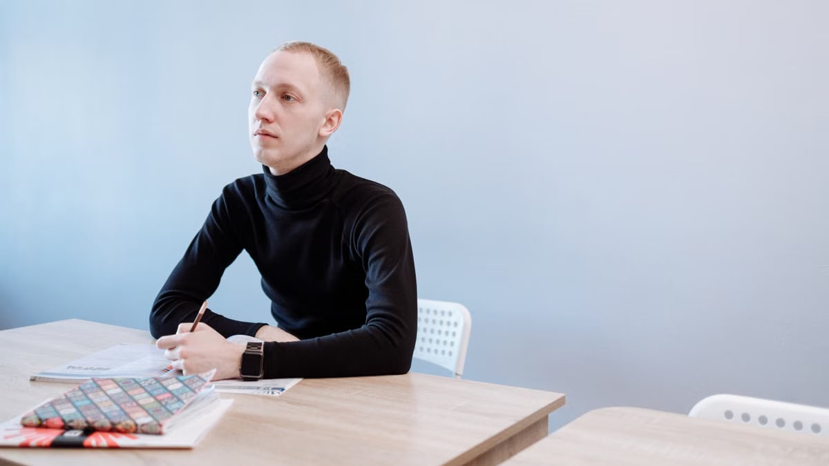 Young man in a black turtleneck writing at a desk, contemplating ideas.